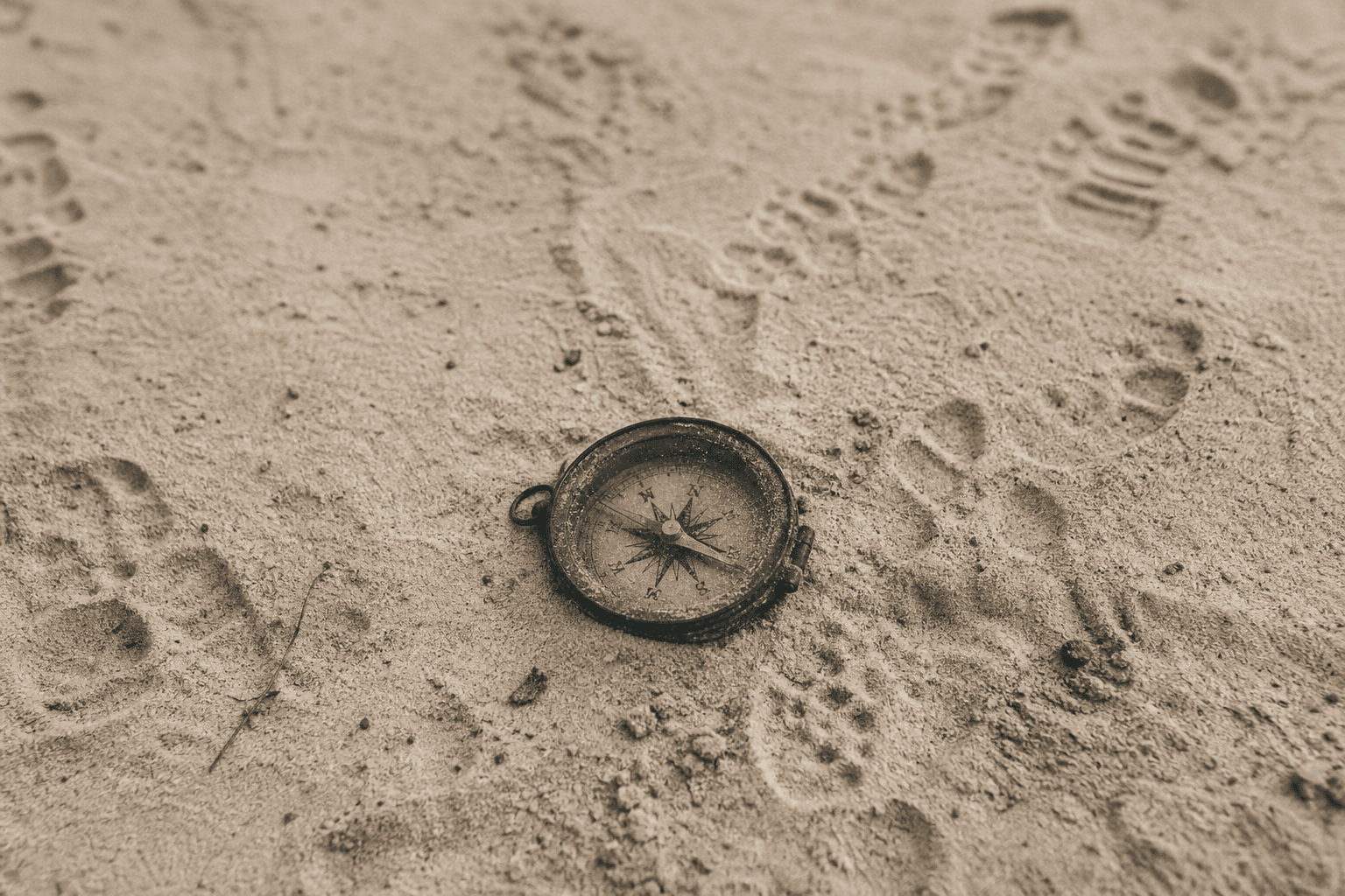 A weathered compass half-buried in dry, textured sand with subtle footprints trailing off in multiple directions around it, photographed from above in soft, diffused natural light, editorial photography style with muted warm tones and visible film grain, color palette of warm off-white sand, olive green patina on the compass brass, warm grey shadows, and deep charcoal details, shallow depth of field creating a quiet and introspective atmosphere, the compass needle slightly askew between directions, landscape orientation with wide crop emphasizing the surrounding emptiness and the gentle chaos of overlapping tracks in the sand